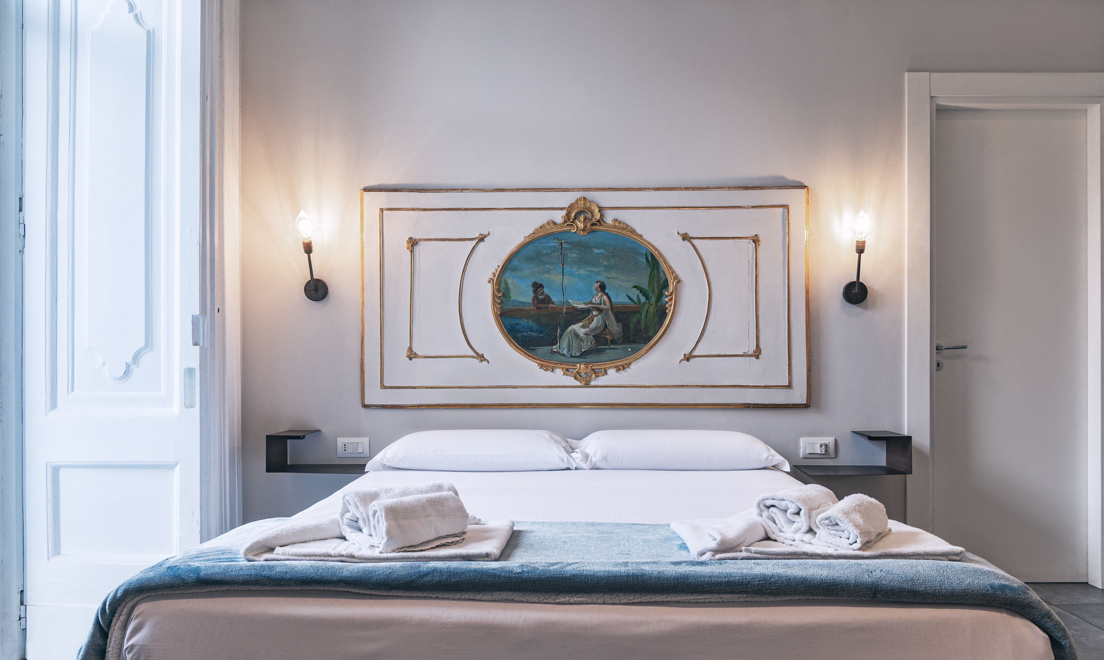 A hotel room with a bed, two wall sconces, a painting, and a door.