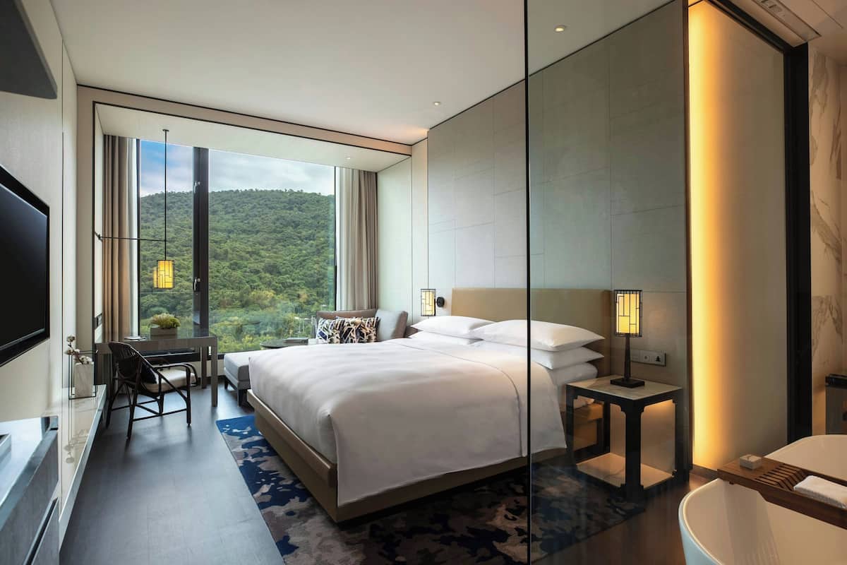 Habitación de hotel moderna con una cama grande, un escritorio y vistas a una colina boscosa.