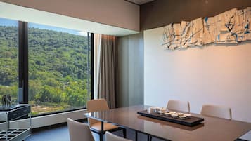 A modern conference room with a large table, white chairs, and a view of a forested hillside through large windows.
