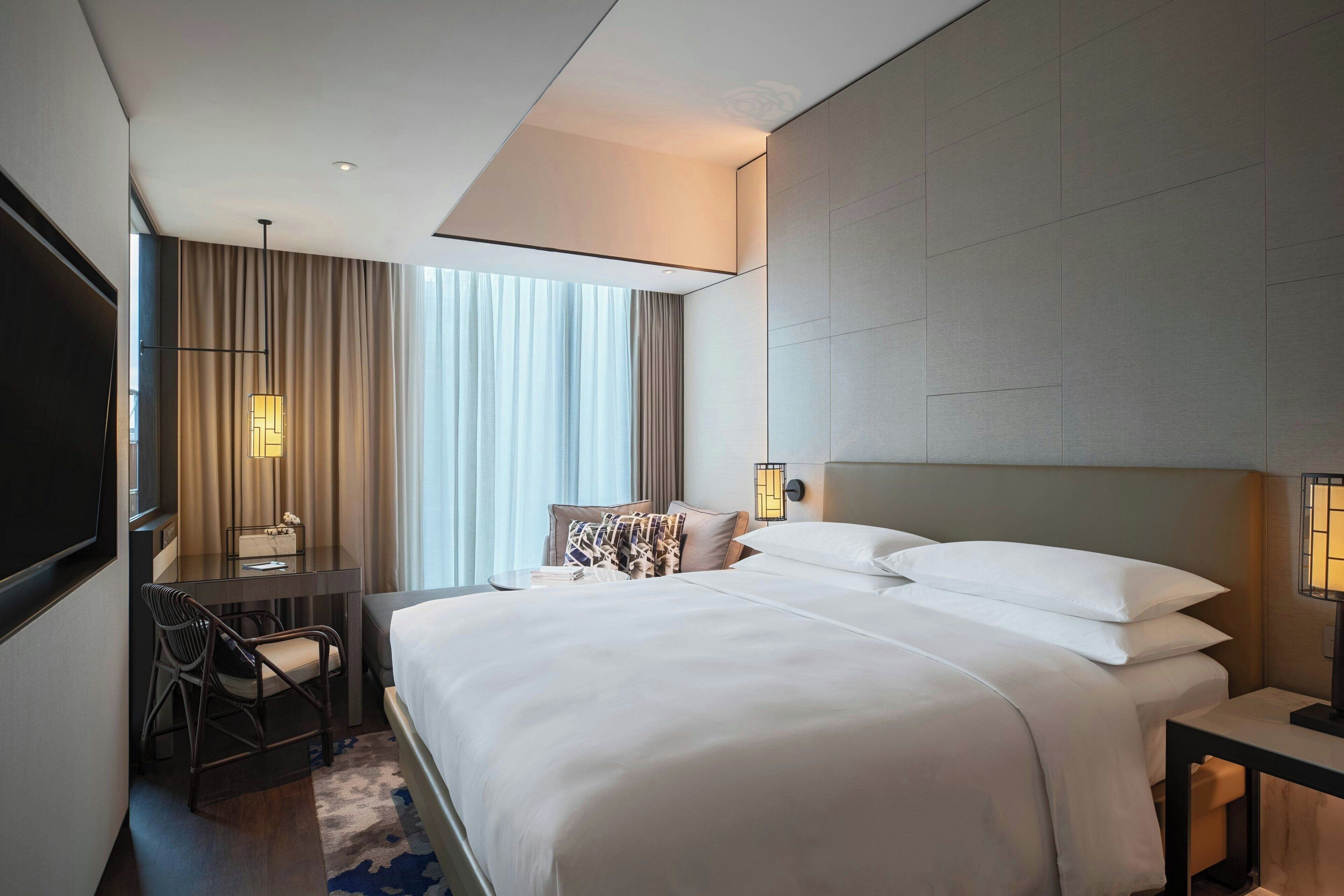 A modern hotel room with a large bed, a desk, and a chair. There is a window with curtains and a lamp on the desk.