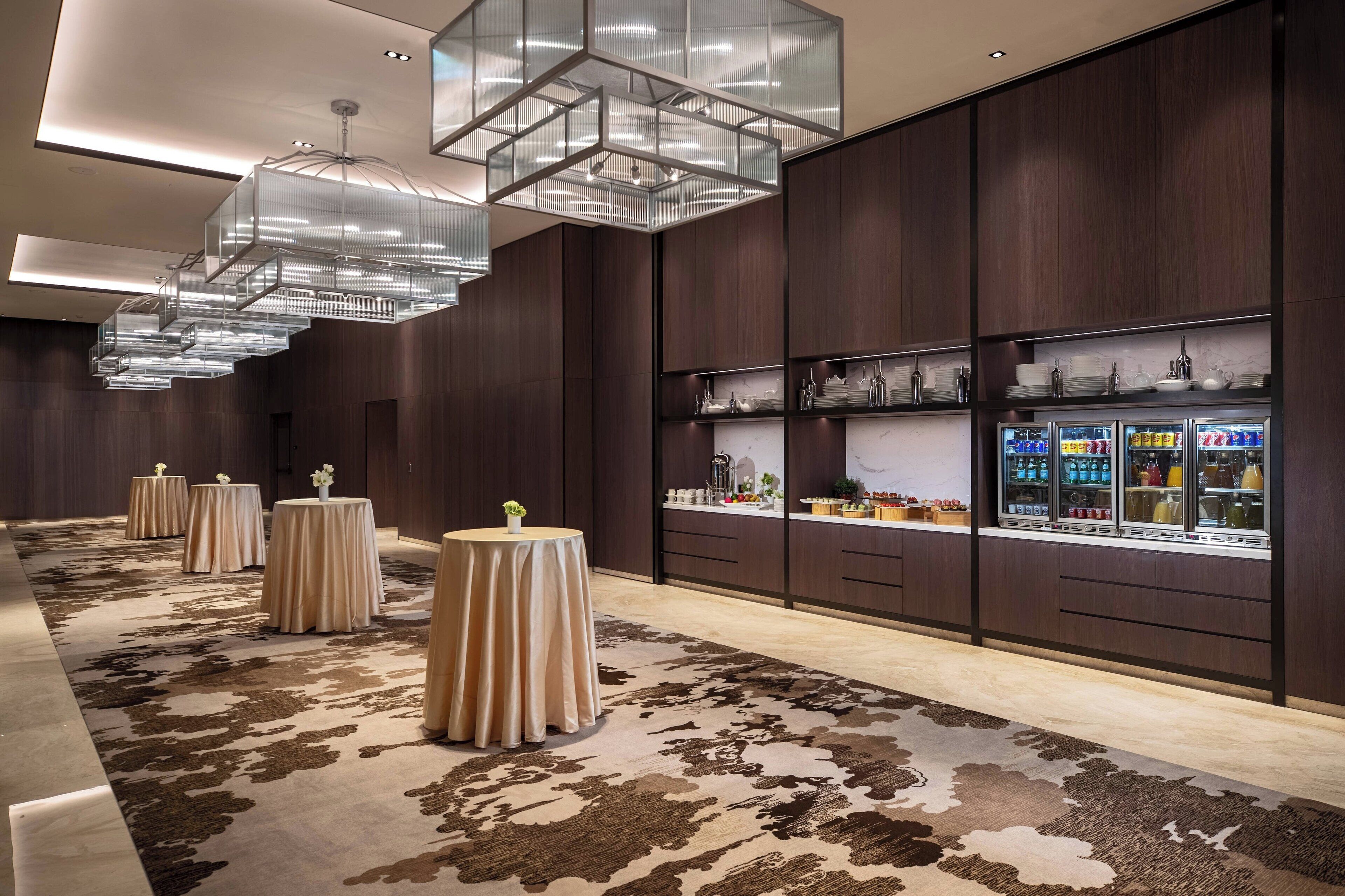 A modern hotel lobby with a patterned carpet, round tables with tablecloths, and a display of glassware and bottles.