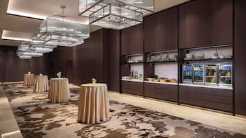 A modern hotel lobby with a patterned carpet, round tables with tablecloths, and a display of glassware and bottles.