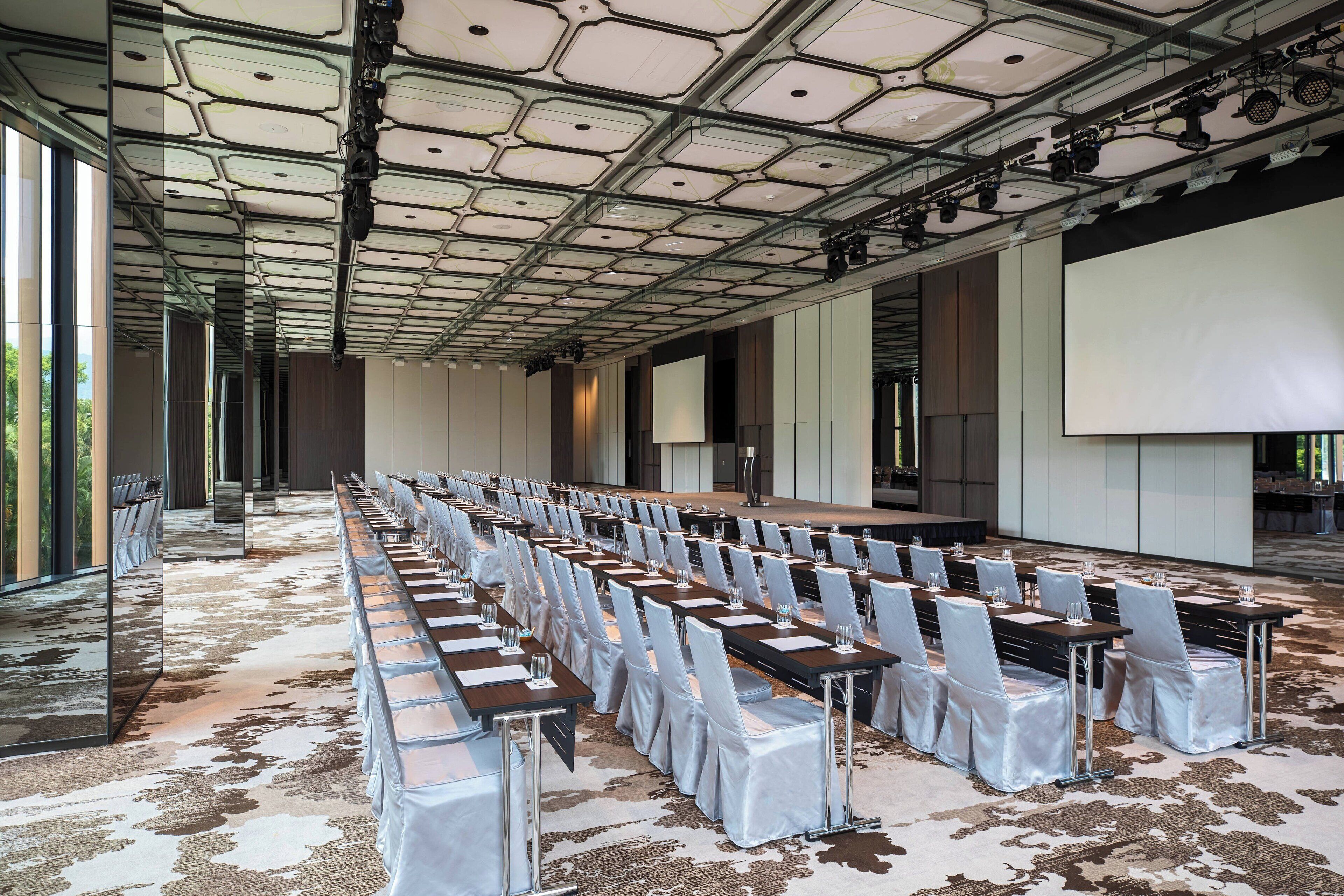 A conference room with rows of tables and chairs, a stage, and a large screen.