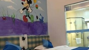 A hotel room with two beds, each with blue and white bedding, a Mickey Mouse mural on the wall, and a window with a view of the room's interior.