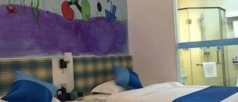 Habitación de hotel con dos camas, cada una con ropa de cama azul y blanca, un mural de Mickey Mouse en la pared y una ventana con vista al interior de la habitación.