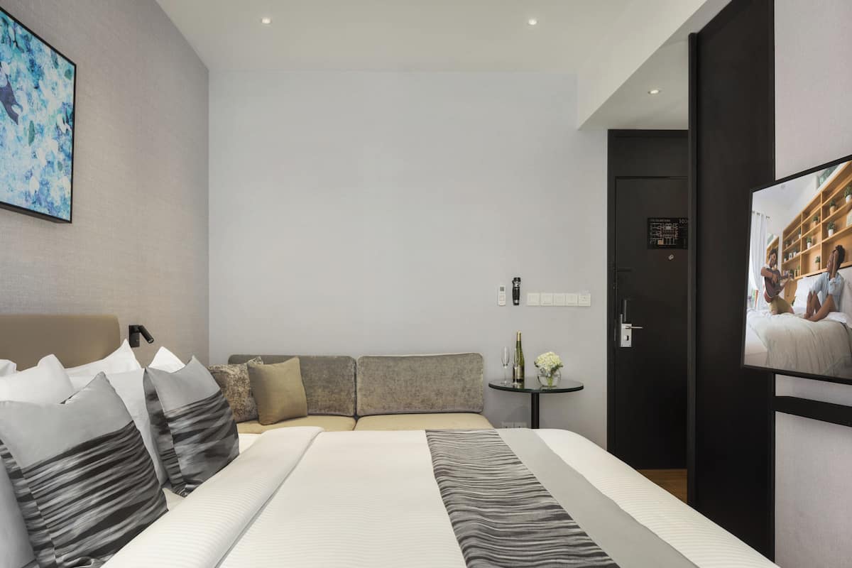 Ein modernes Hotelzimmer mit einem großen Bett, einer Sitzecke, einem kleinen Tisch mit Flasche und Blumen sowie einem Fernseher an der Wand.