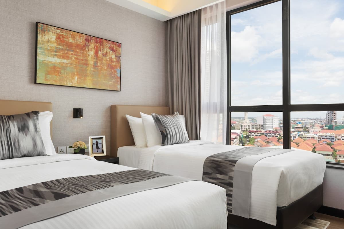 Ein Hotelzimmer mit zwei Betten, einem großen Fenster mit Blick auf die Stadt, einem Gemälde an der Wand und einem Nachttisch mit Blumenvase.
