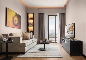 A modern living room with a sofa, coffee table, and TV stand. There is a large window with curtains, a painting on the wall, and a bookshelf.