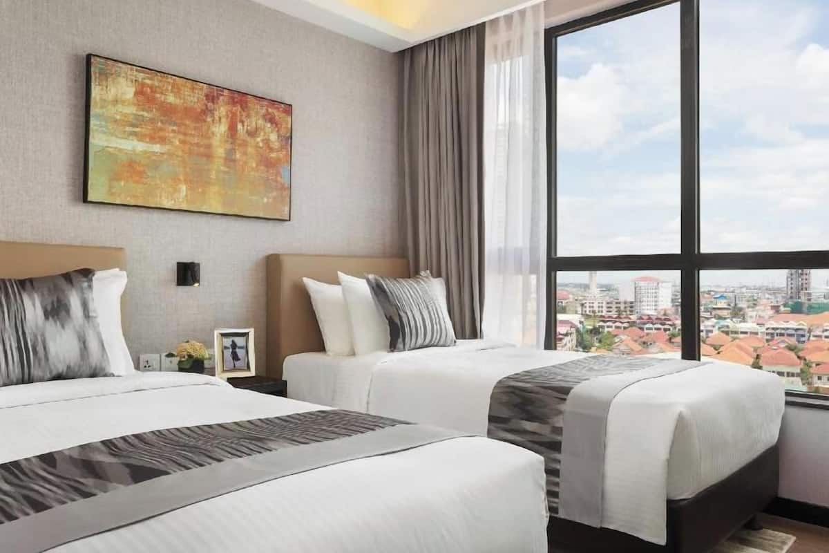 Ein Hotelzimmer mit zwei Betten, einem großen Fenster mit Blick auf die Stadt, einem Bild an der Wand und einem Nachttisch mit einer Lampe.