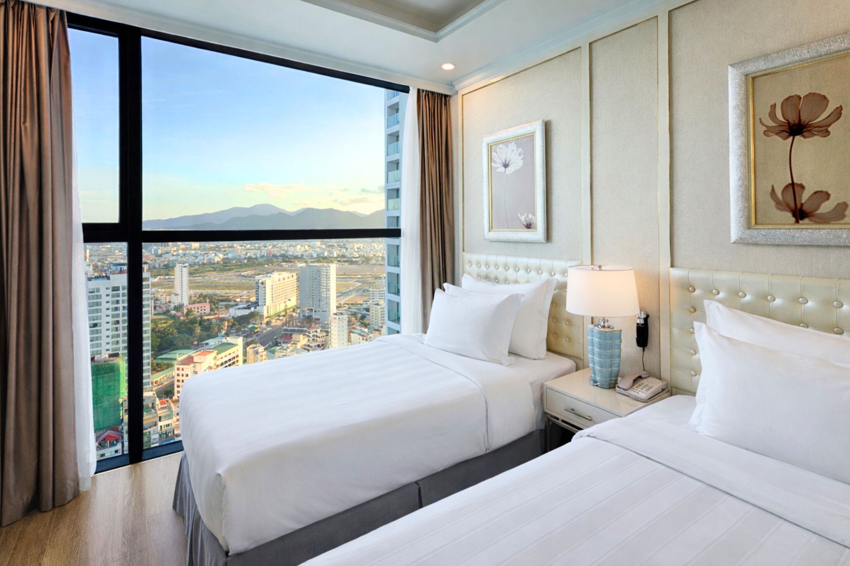 Ein Hotelzimmer mit zwei Betten, einem großen Fenster mit Blick auf die Stadt, einem Nachttisch mit Lampe und einem gerahmten Kunstwerk an der Wand.