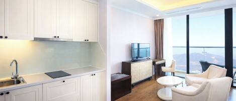 A modern kitchen with white cabinets, a built-in stove, and a sink. There is a seating area with a round table and two chairs. A flat-screen TV is mounted on the wall.