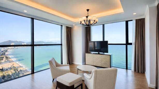 Une chambre d’hôtel moderne dotée d’une grande fenêtre offrant une vue sur l’océan et le paysage urbain, d’un téléviseur à écran plat et d’un coin salon avec une petite table.