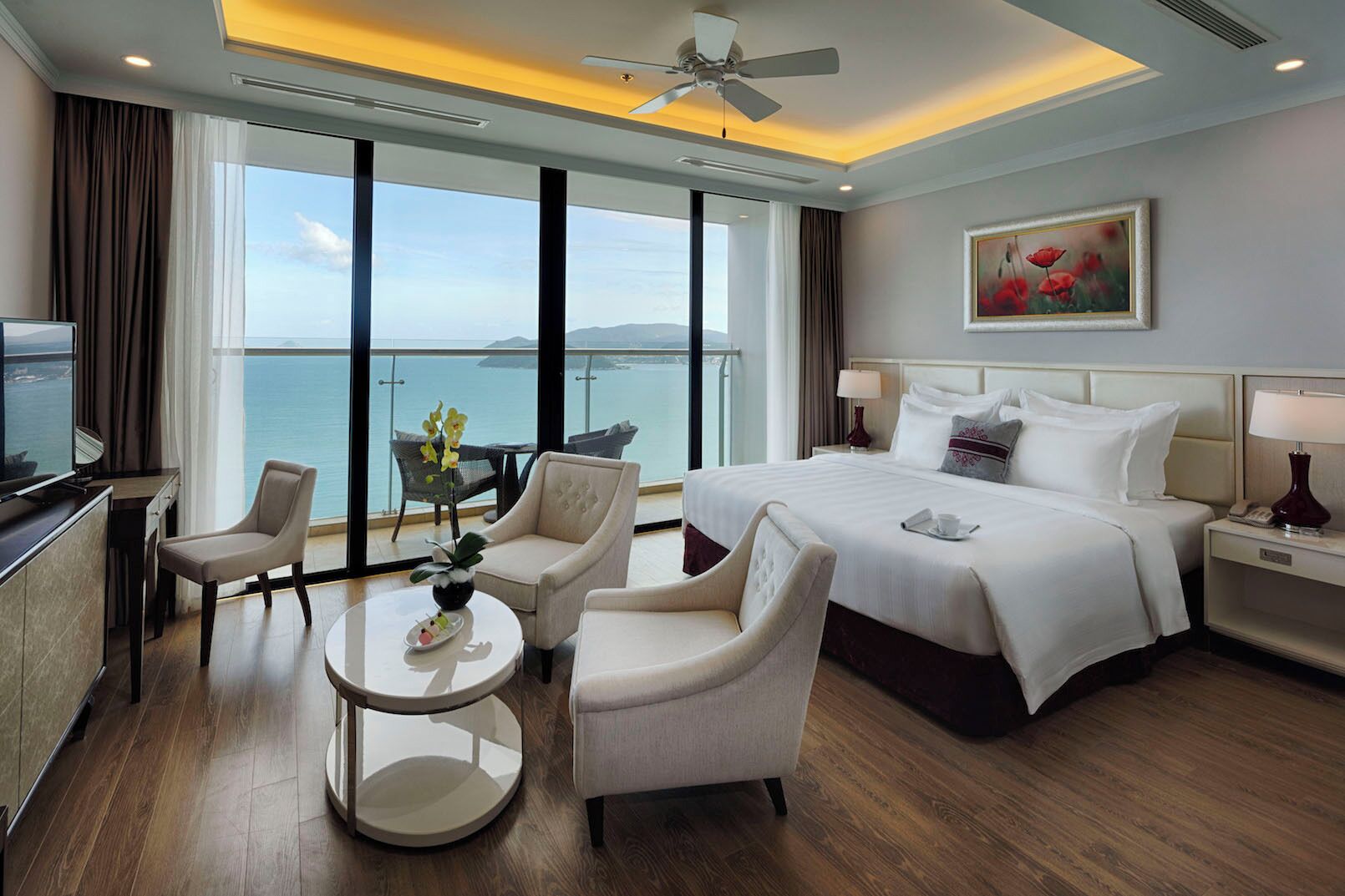 A hotel room with a bed, two armchairs, a coffee table, a desk with a TV, and a large window with a view of the sea.