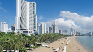 A beachfront hotel with high-rise buildings in the background, a sandy beach, and palm trees.