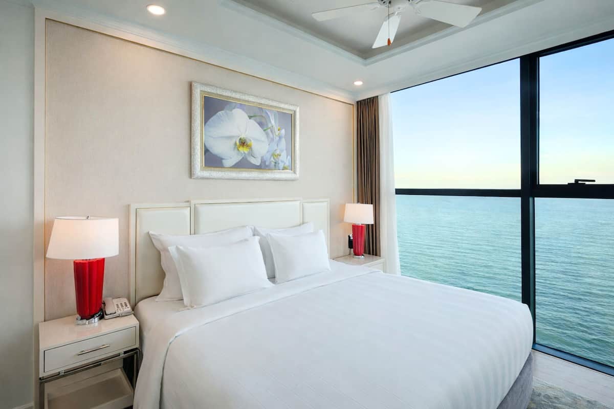 Een hotelkamer met een groot bed, twee nachtkastjes met lampen, een plafondventilator, een groot raam met uitzicht op het water, en een ingelijste foto aan de muur.