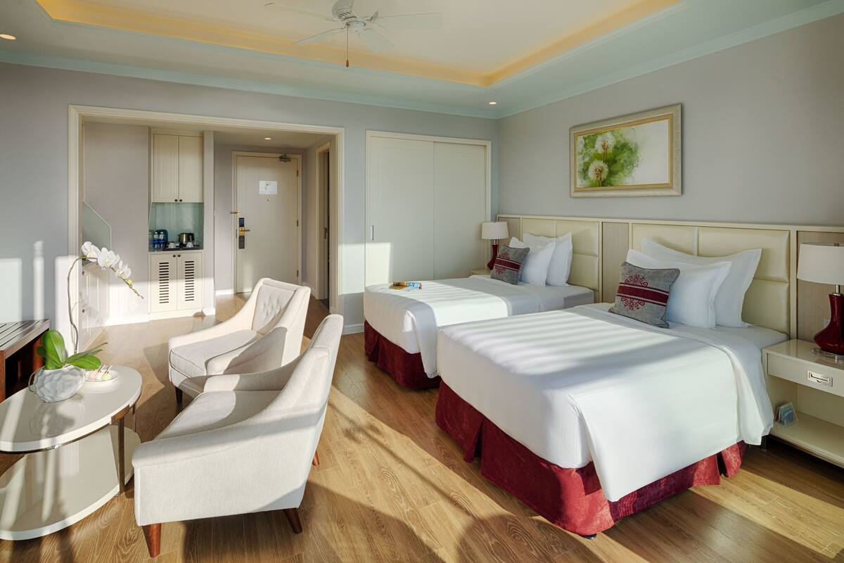 Een hotelkamer met twee bedden, een zithoek met een stoel en een tafel, een plafondventilator en een schilderij aan de muur.