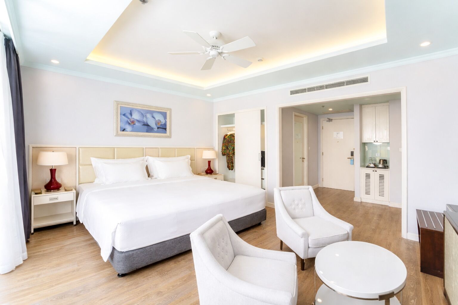 A hotel room with a large bed, two bedside tables with lamps, a ceiling fan, a dining area with a table and chairs, and a kitchenette.