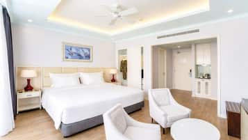 A hotel room with a large bed, two bedside tables with lamps, a ceiling fan, a dining area with a table and chairs, and a kitchenette.