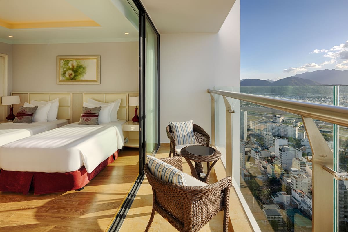 Een hotelkamer met twee bedden, een balkon met rieten meubels en uitzicht op de stad.