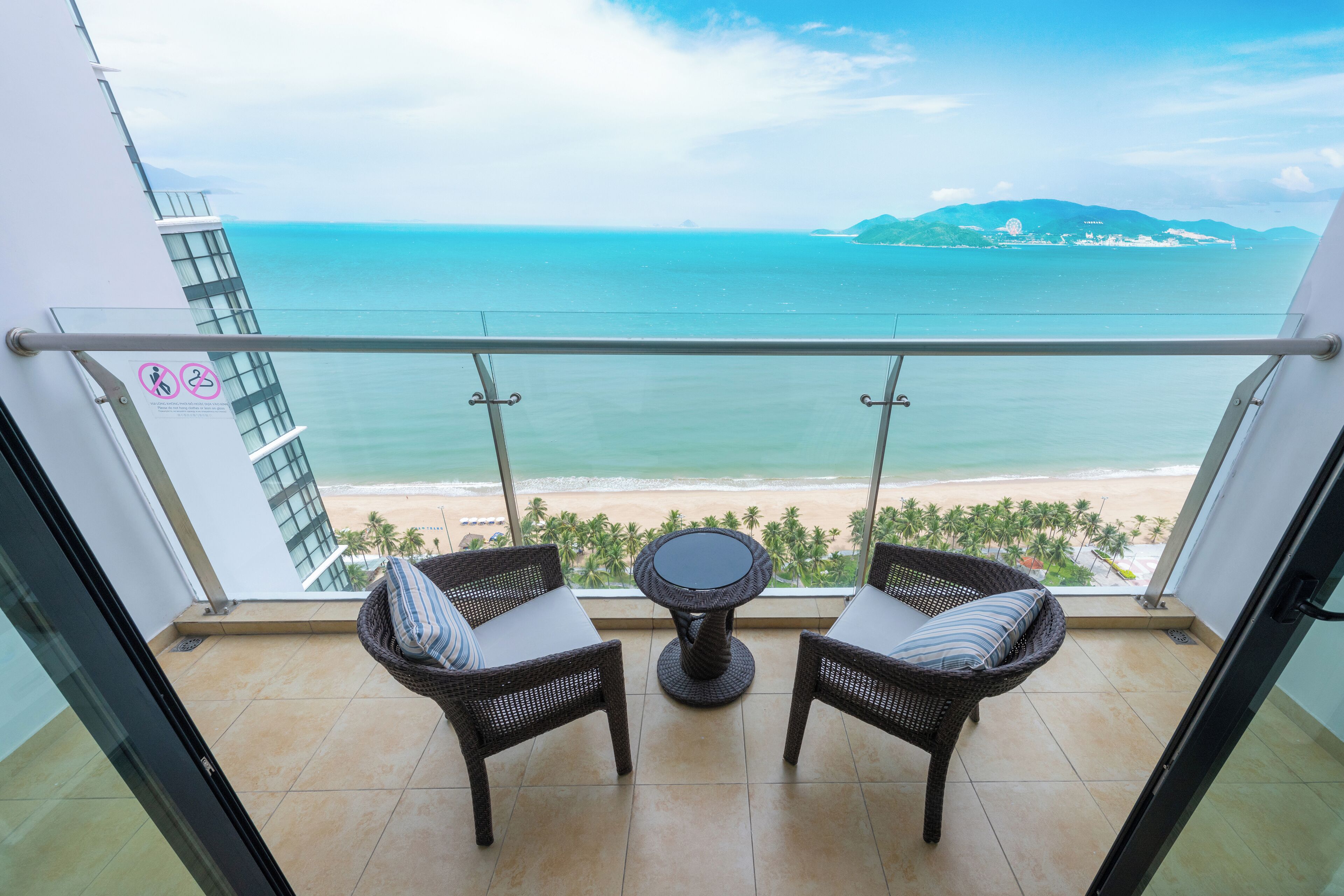 A balcony with two wicker chairs and a small table, overlooking a beach and ocean.
