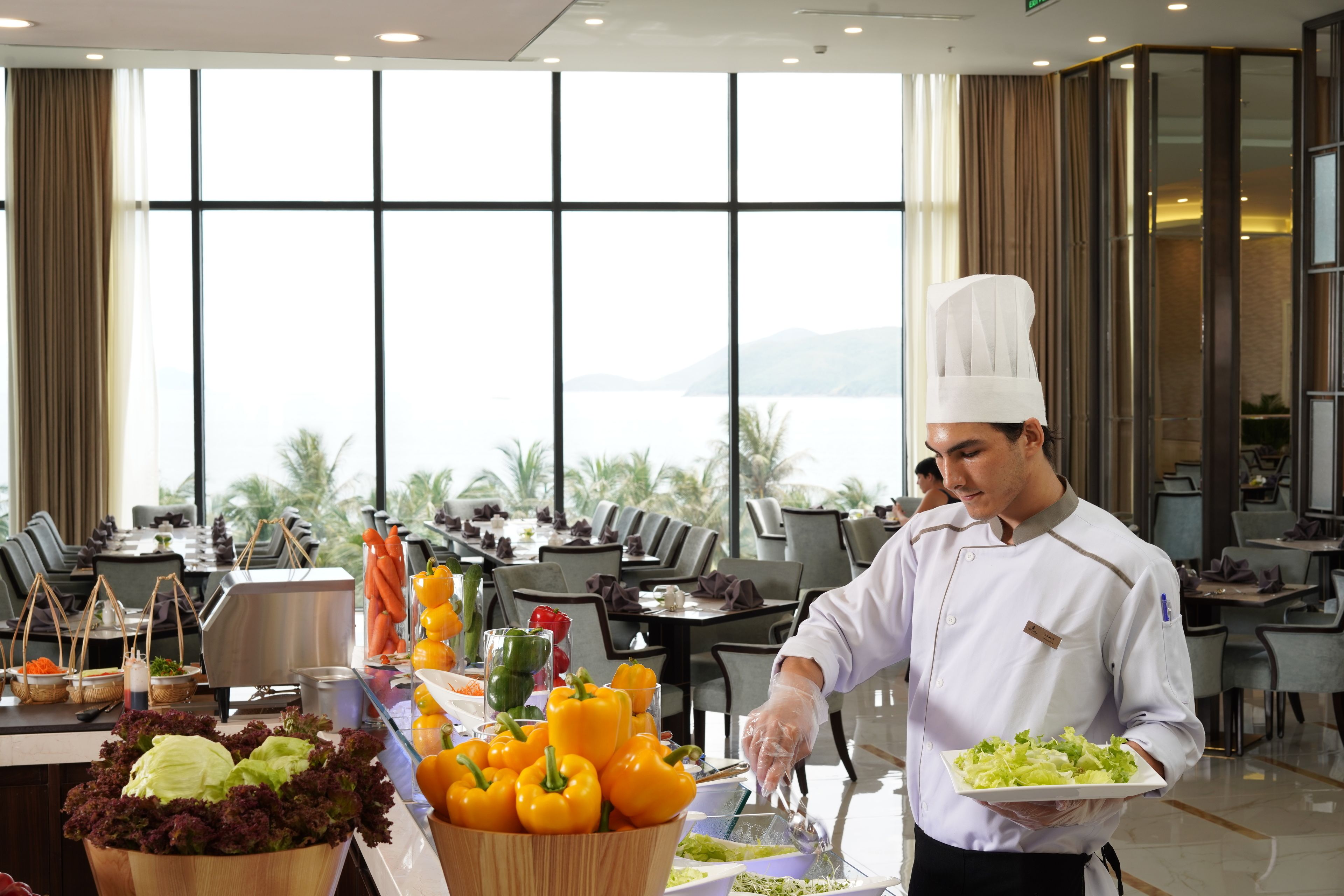 Un chef con uniforme blanco y gorro de cocinero sirve un plato de ensalada. Al fondo, hay mesas con sillas, un bufé con diversas verduras y vistas a montañas a través de amplias ventanas.