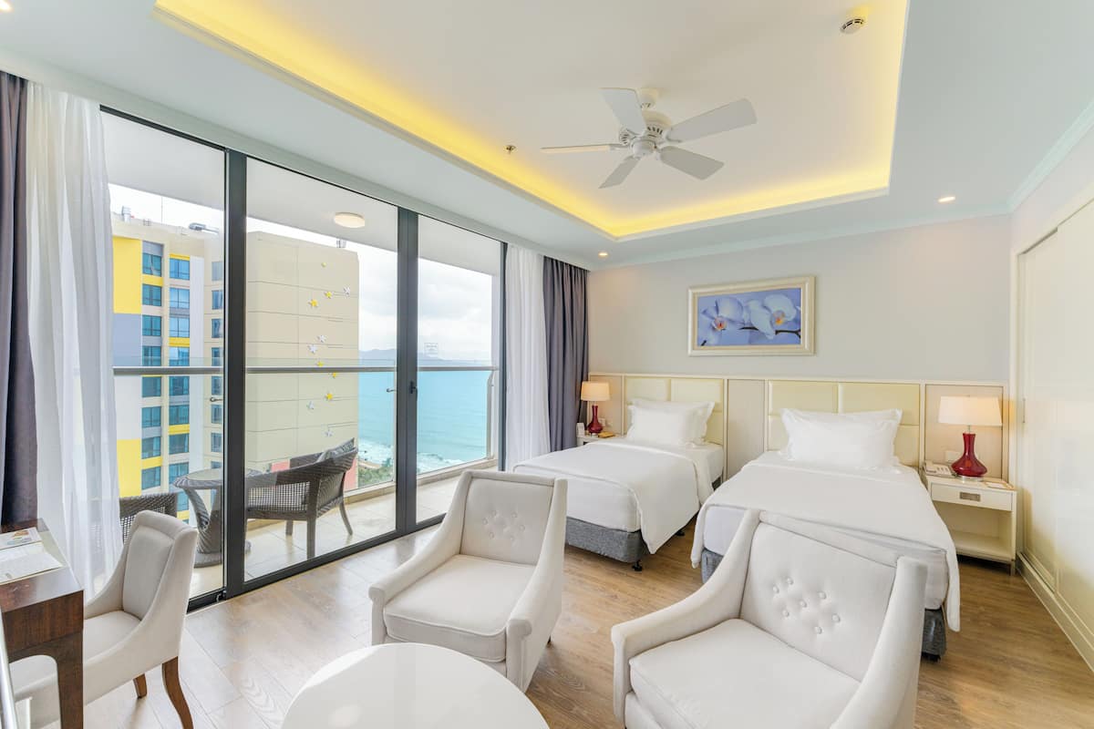 Hotelkamer met twee bedden, een bureau, een stoel, een balkon met uitzicht op gebouwen en de zee, en een plafondventilator.