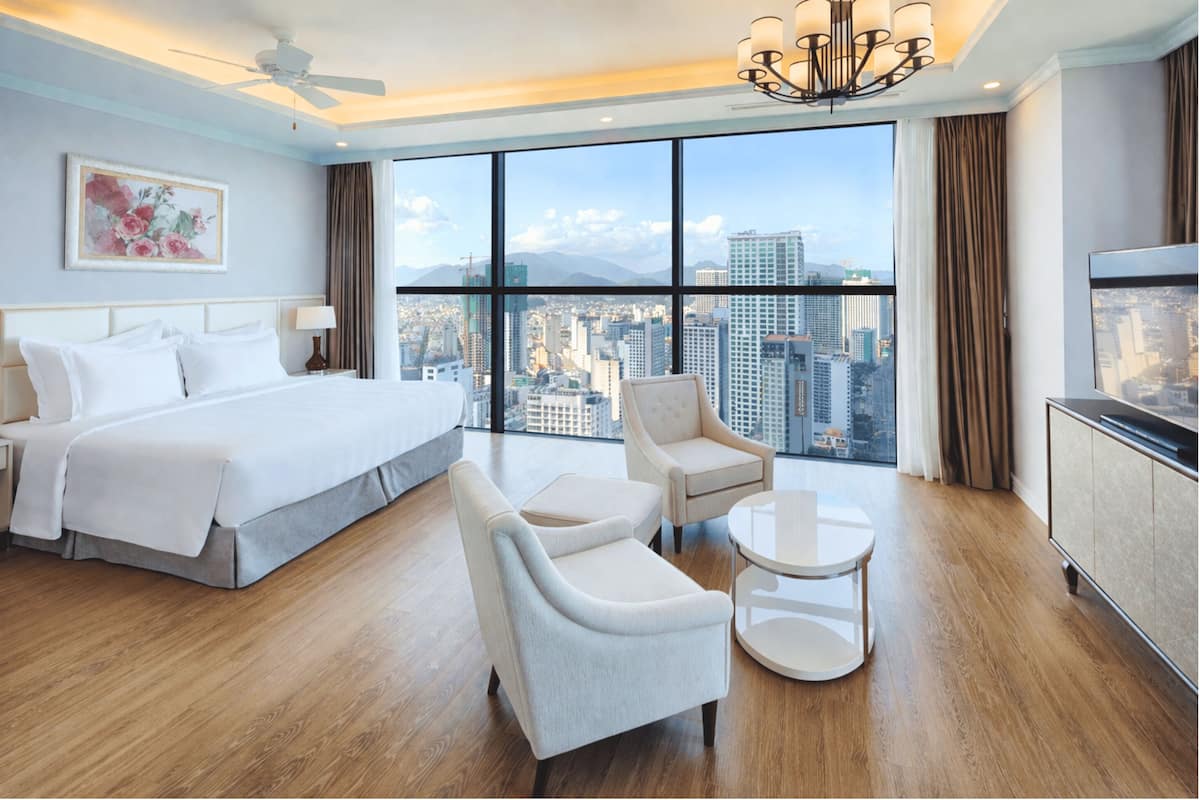 Hotelkamer met een groot bed, twee fauteuils, een bijzettafeltje, een plafondventilator en uitzicht op de stad door grote ramen.