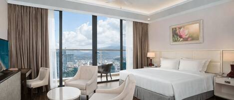 A hotel room with a bed, two armchairs, a small table, a desk, a ceiling fan, a painting, and a view of the city.