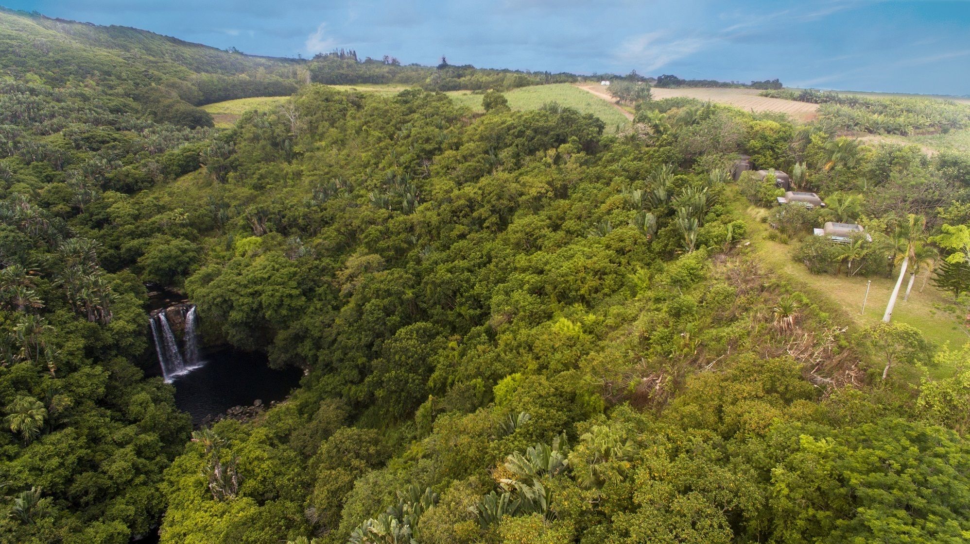 An aerial view of a lush, green landscape with a waterfall and a small building surrounded by dense vegetation.