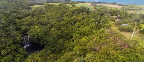 An aerial view of a lush, green landscape with a waterfall and a small building surrounded by dense vegetation.