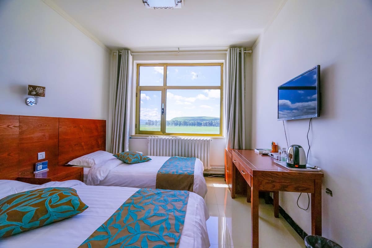 Une chambre d’hôtel avec deux lits, une tête de lit en bois, un bureau avec une bouilloire, une télévision et une fenêtre donnant sur la campagne.