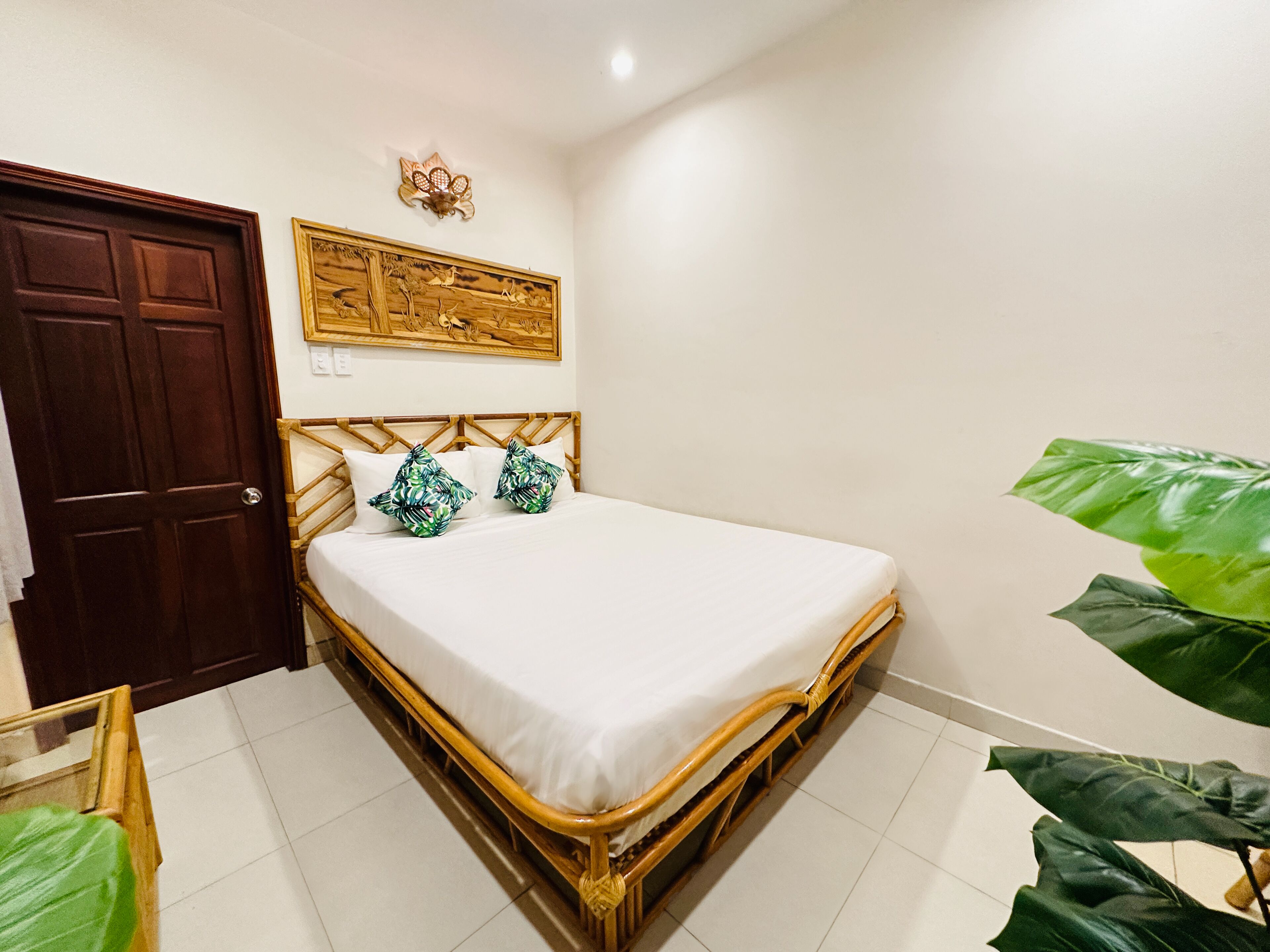 A room with a wooden bed, white bedding, a wooden headboard, a door, a wall-mounted light fixture, and a large green plant.