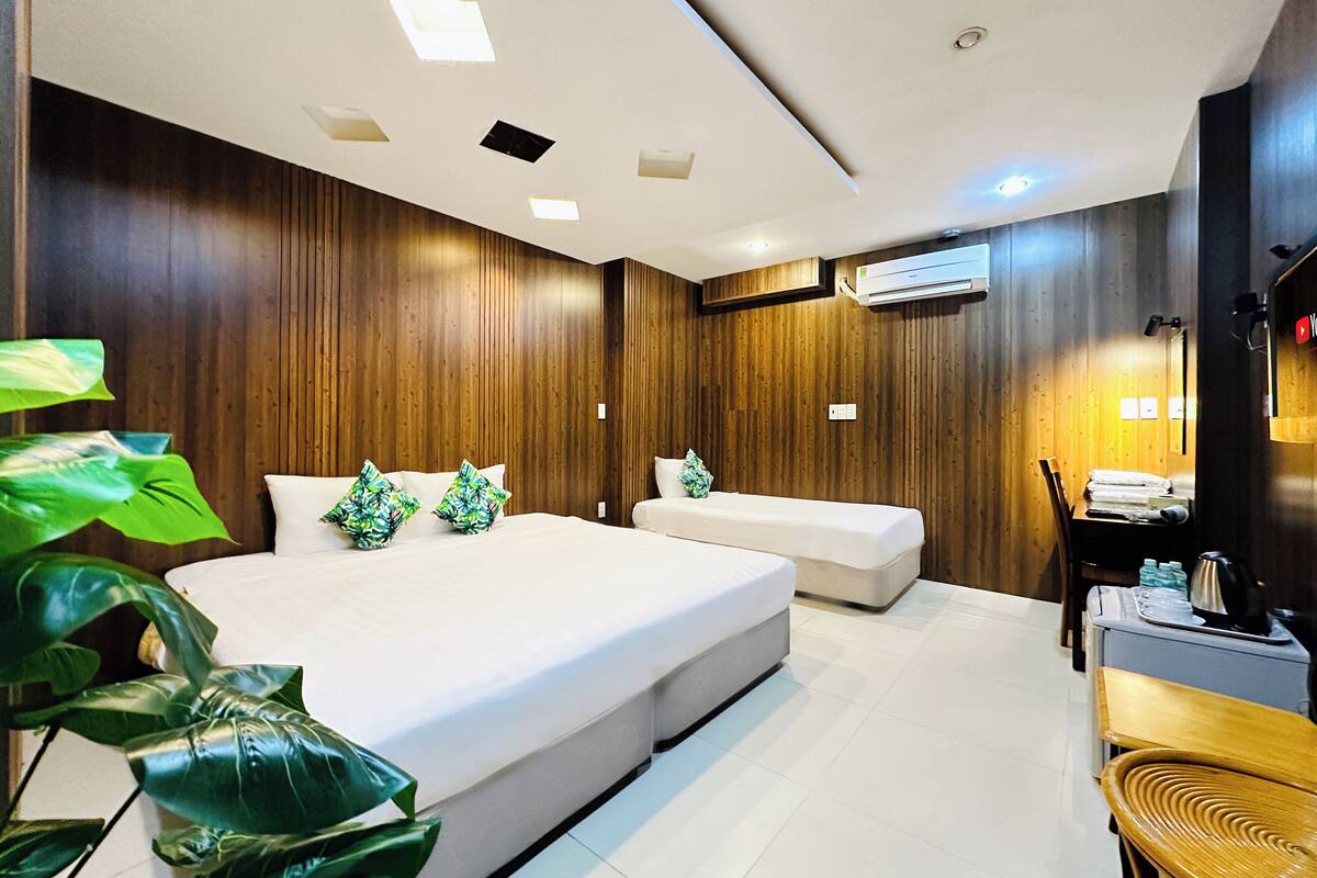 Ein modernes Hotelzimmer mit einem großen Bett, einem Einzelbett, einem Schreibtisch mit Stuhl und einer großen Zimmerpflanze.