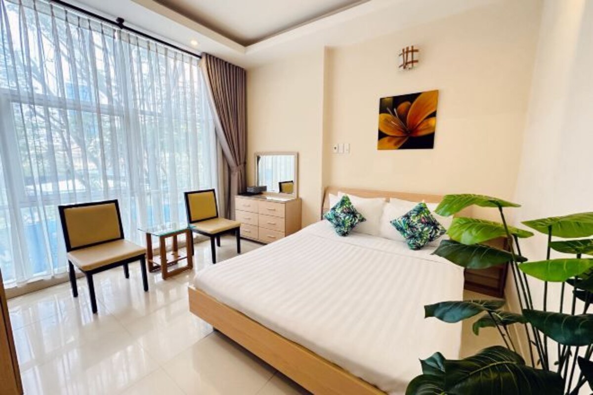 Une chambre d’hôtel moderne avec un grand lit, un bureau et une chaise. Il y a une fenêtre avec des rideaux légers et une plante près du lit.