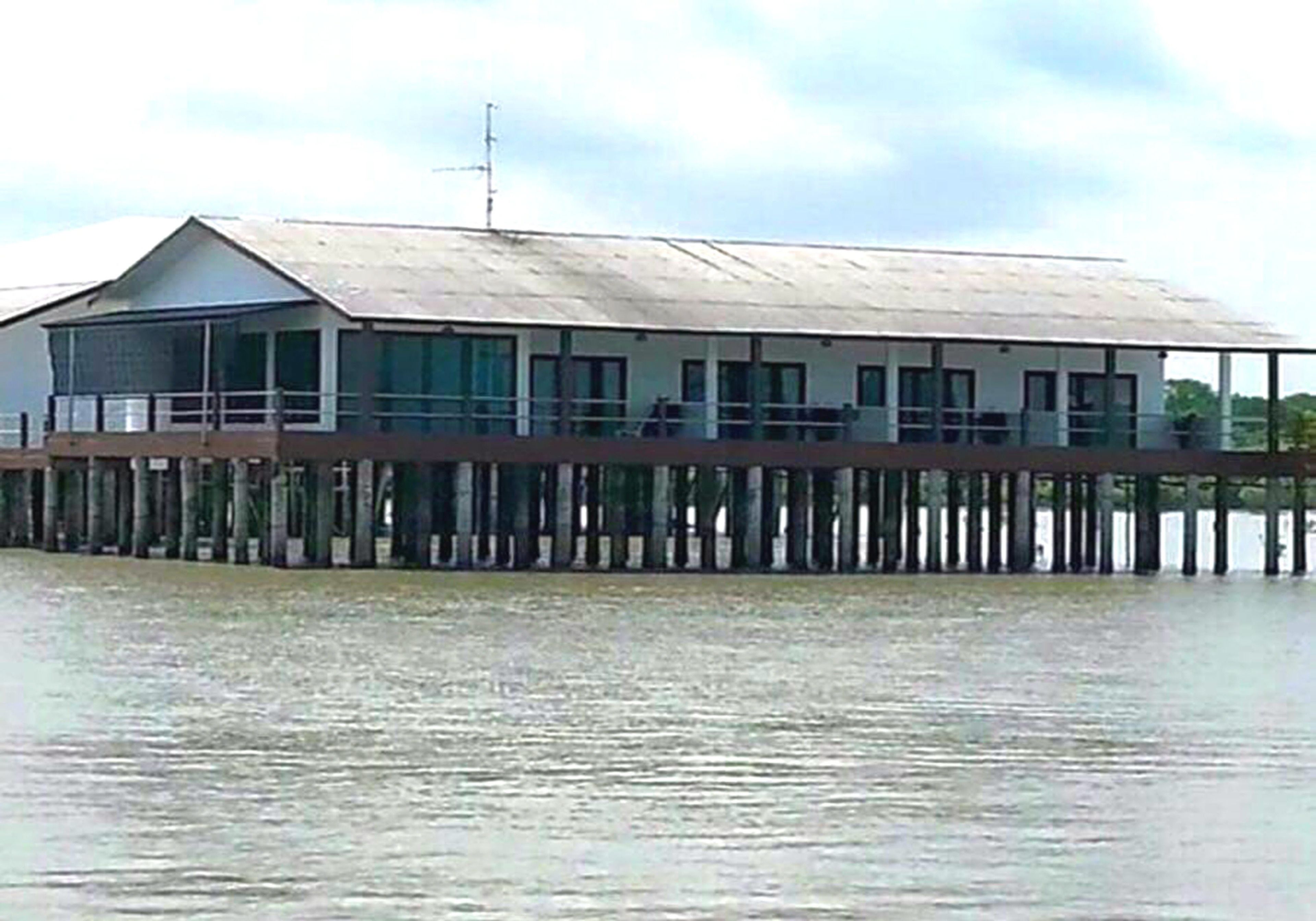 A building with a flat roof, large windows, and a balcony. It is supported by wooden stilts over water.