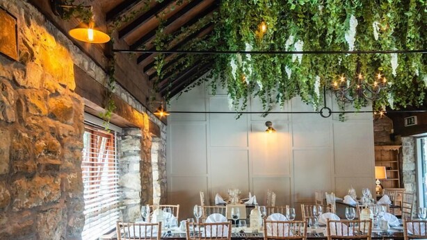A restaurant with rustic stone walls, wooden beams, and hanging greenery. Tables are set with white tablecloths, silverware, and wine glasses. - Tully Mill Cottages (Enniskillen)