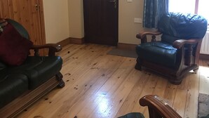 A room with a wooden floor, a dark wooden door, a green leather sofa, a wooden armchair, and a window with curtains. - Tully Mill Cottages (Enniskillen)