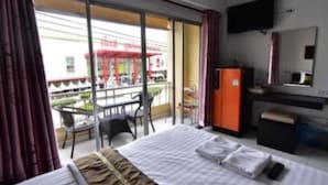 A hotel room with a bed, a desk, a chair, a television, and a view of an outdoor area with a building and a red umbrella. - CK Residence (Pattaya)