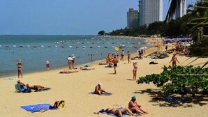 A beach with people sunbathing and swimming, palm trees, and a cityscape in the background. - CK Residence (Pattaya)