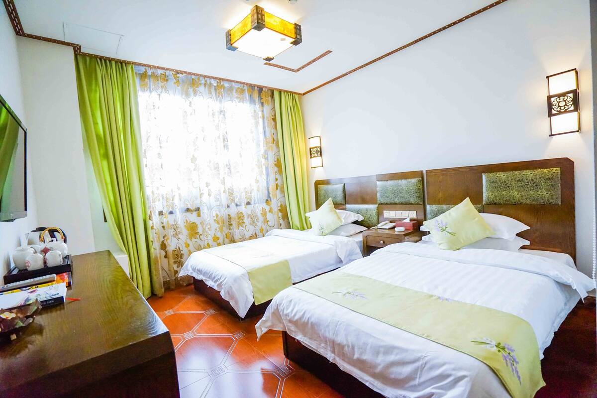 Quarto de hotel com duas camas, uma escrivaninha de madeira, televisão e janela com cortinas.