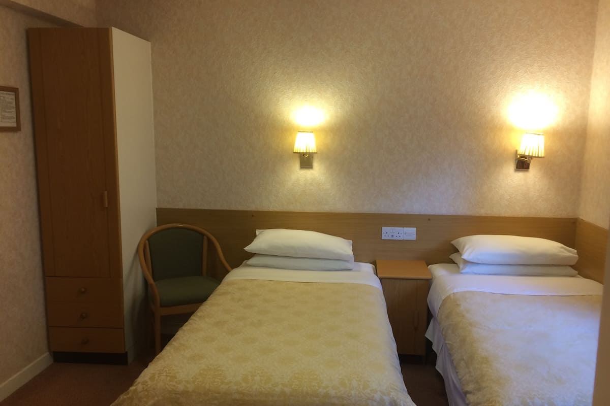 A hotel room with two single beds, a wooden wardrobe, a green chair, and two wall-mounted lamps.