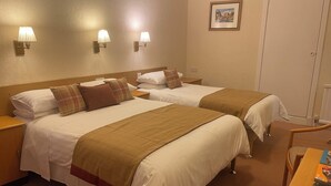 Habitación de hotel con dos camas, cada una con ropa blanca y mantas marrones, un cabecero de madera y una mesita de noche con una lámpara.