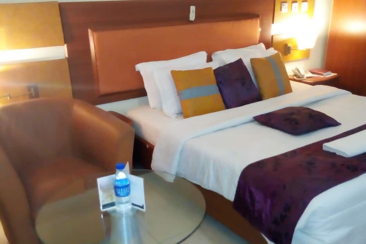 Quarto de hotel com uma cama, uma cadeira, uma mesinha com uma garrafa de água e um criado-mudo com abajur.