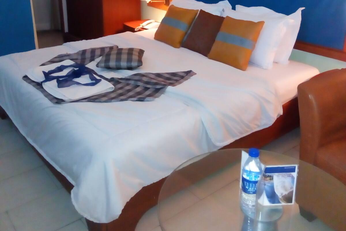 Quarto de hotel com uma cama bem arrumada, uma mesa de cabeceira com uma garrafa de água e um folheto, e uma poltrona marrom.