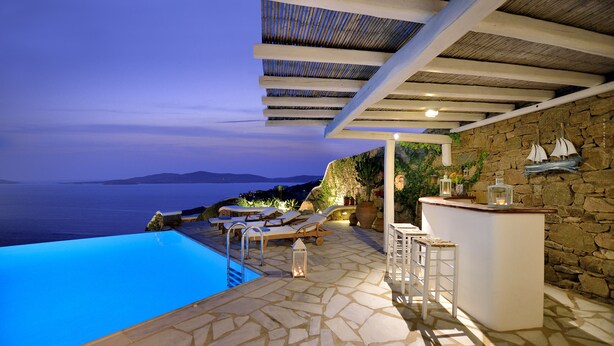 Una piscina infinita con vista al mar, sillas de descanso, zona de bar y suelo de piedra.