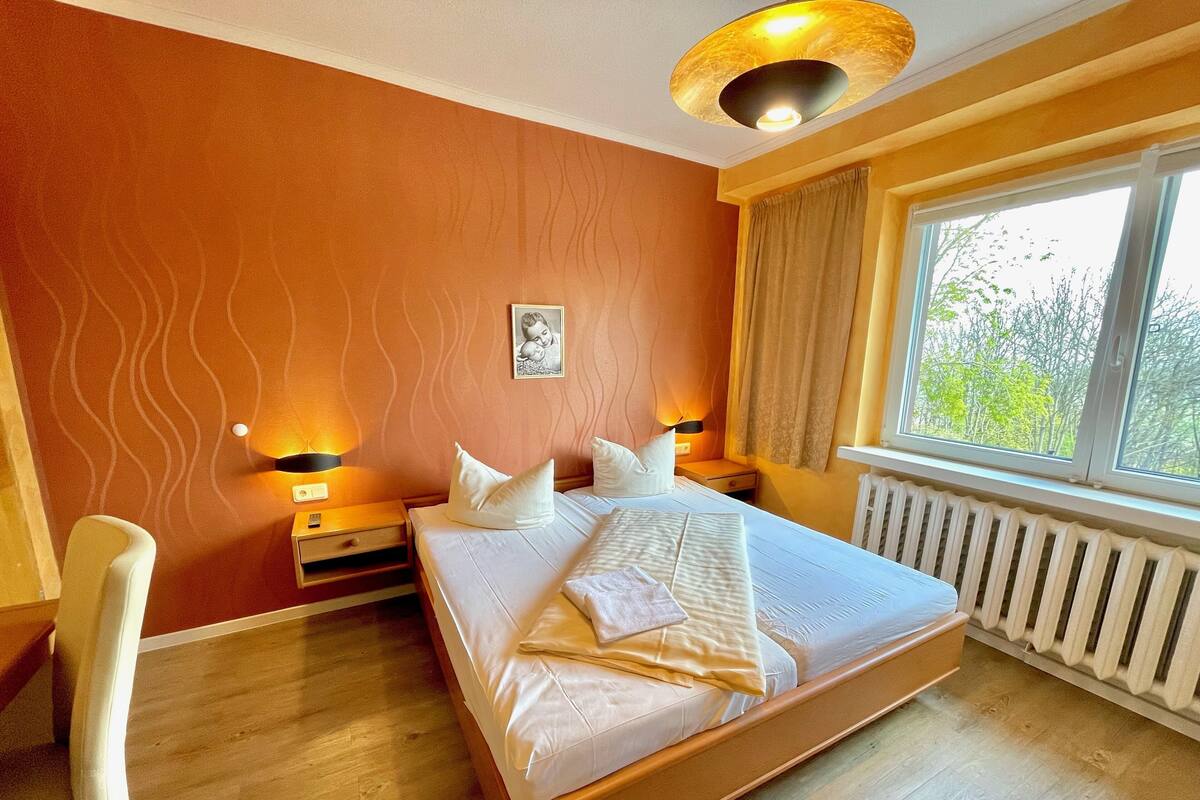 Una camera d'albergo con un letto grande, due comodini con lampade, una finestra con tende e un termosifone.