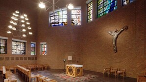 L’intérieur d’une église, avec des bancs disposés en rangées, un autel en bois, des vitraux et un crucifix accroché au mur.