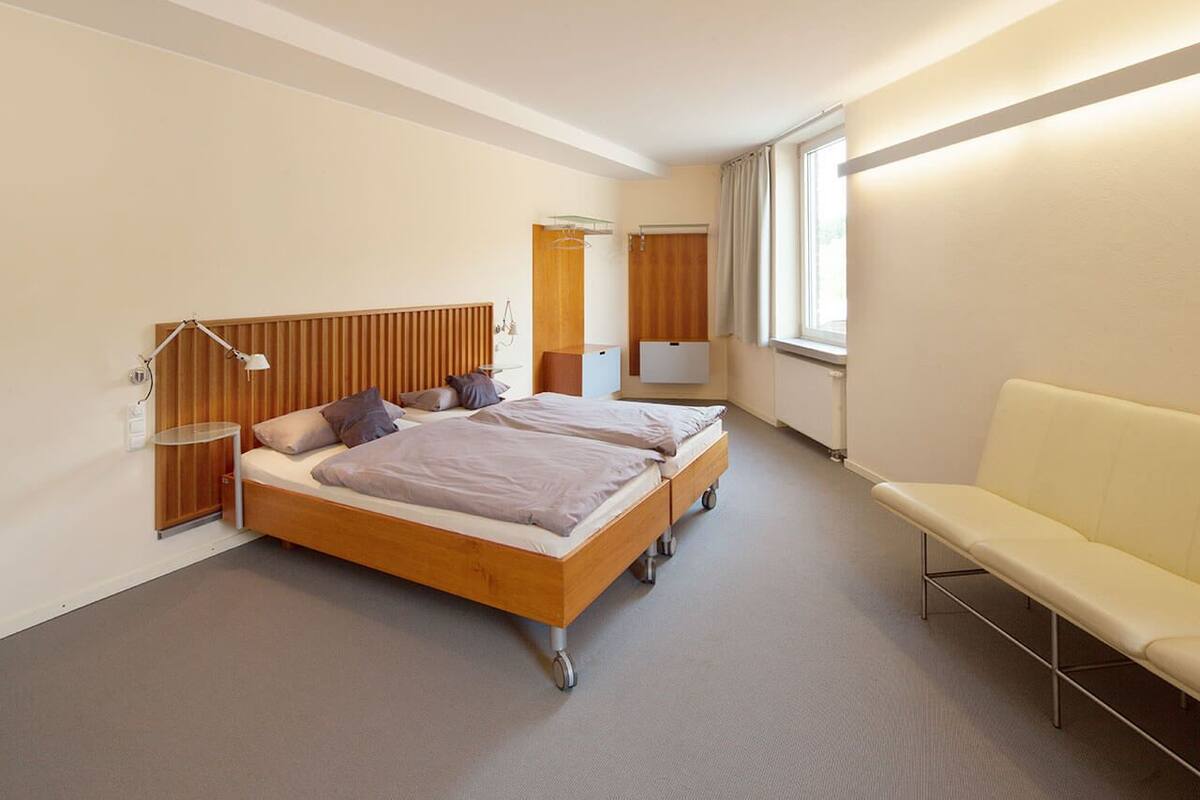 Una habitación de hotel moderna con cama doble, mesita de noche, lámpara, sillón y un ventanal grande.