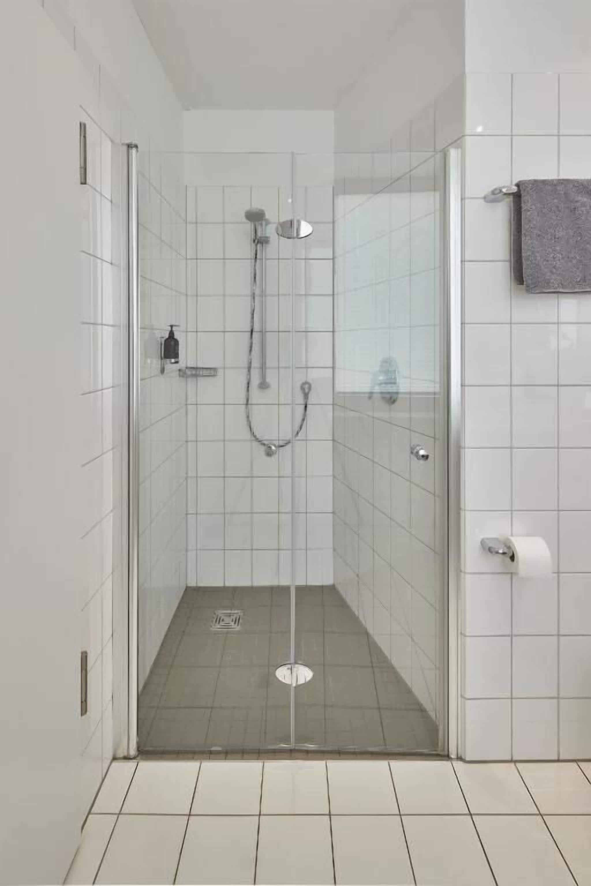 Eine gläserne Duschkabine mit Handbrause, einem wandmontierten Wasserhahn und einem Toilettenpapierhalter auf der rechten Seite.