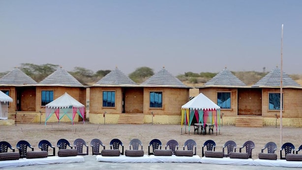 A row of wooden cabins with pitched roofs, each with a small porch and a table and chairs outside. - Shiv Tara Desert Camp (Jaisalmer)